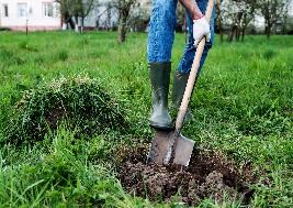 The image shows a person wearing blue jeans, green rubber boots, and white gloves, using a shovel