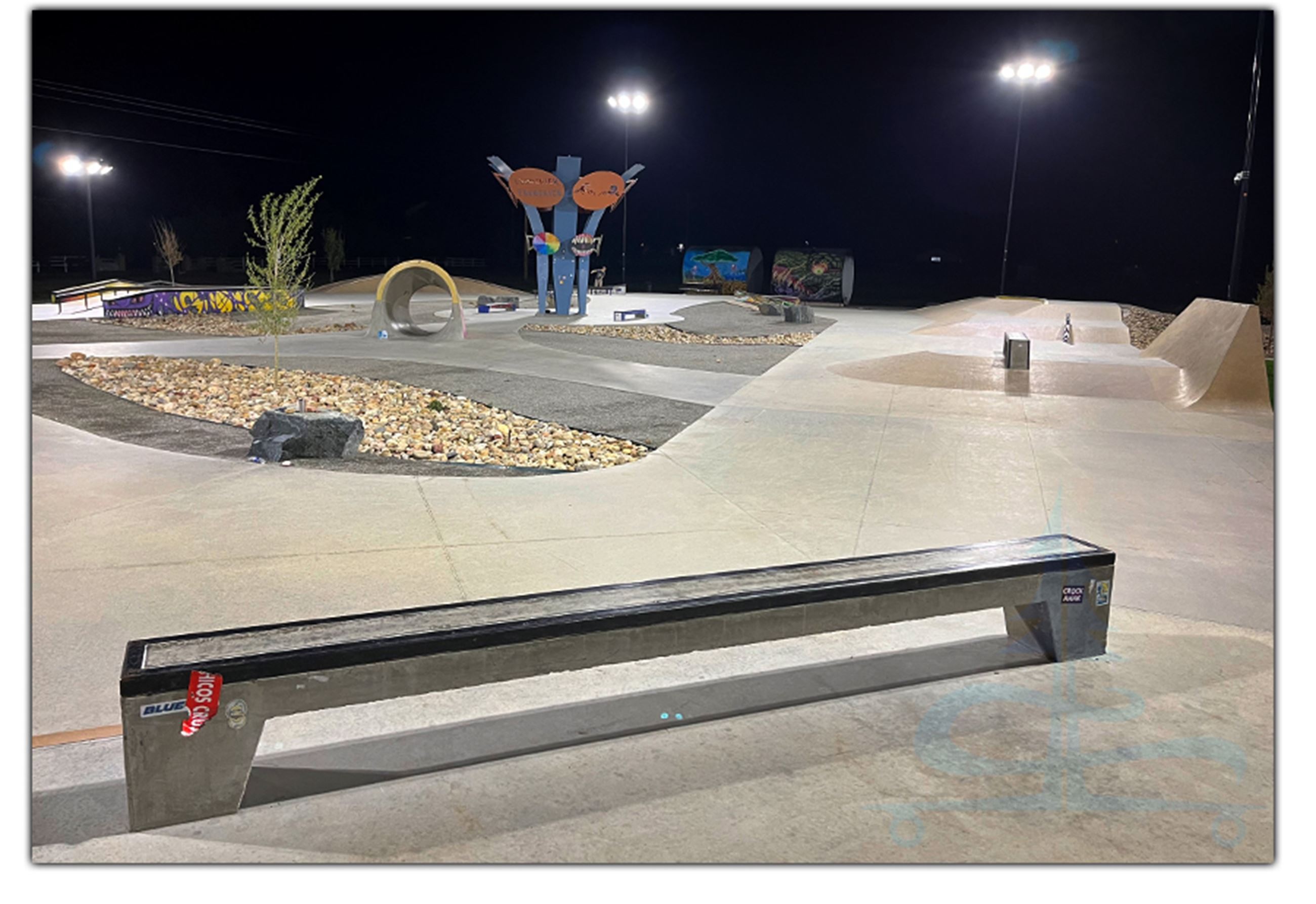 Lit-up skate park at night featuring ramps, rails, and artistic installations.